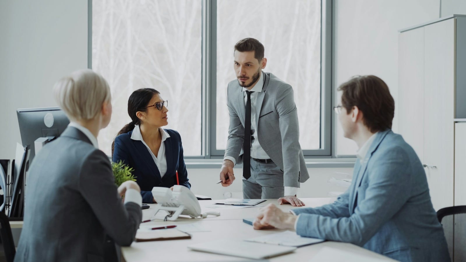 Four people in suits talk around a meeting table; one of them stands and points at some papers.