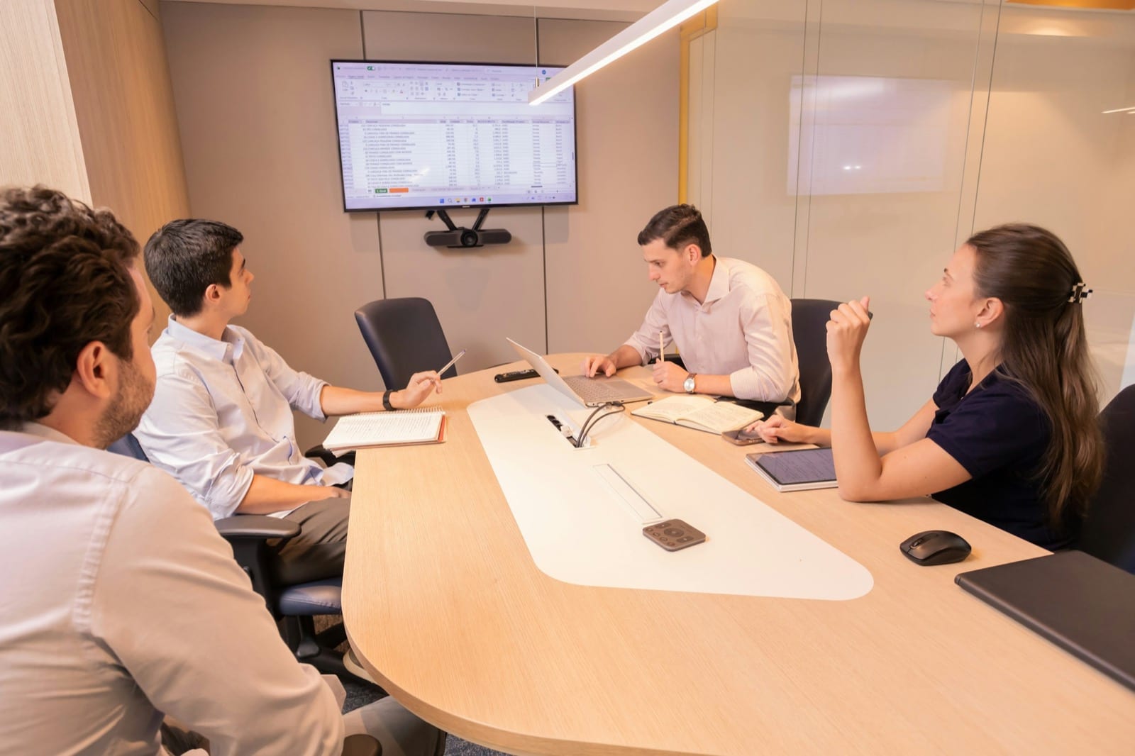 Four people work around a meeting table, facing a screen that shows a spreadsheet.