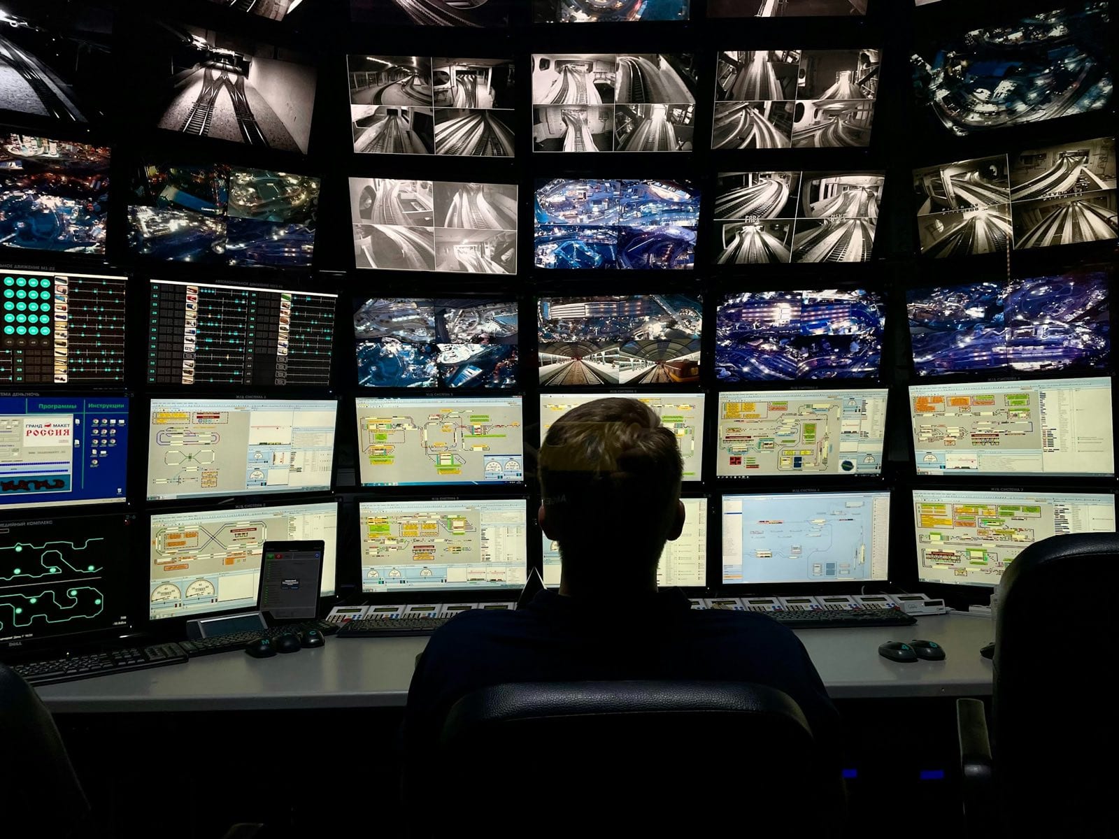 A person, seen from behind, faces a wall of monitors showing camera views and diagrams.