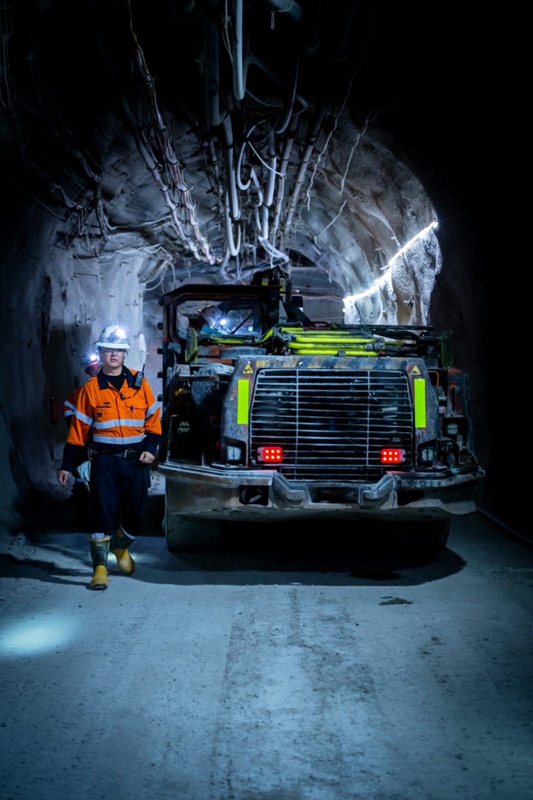 A worker in a helmet with a lamp and high-visibility clothing walks beside a truck in an underground tunnel.