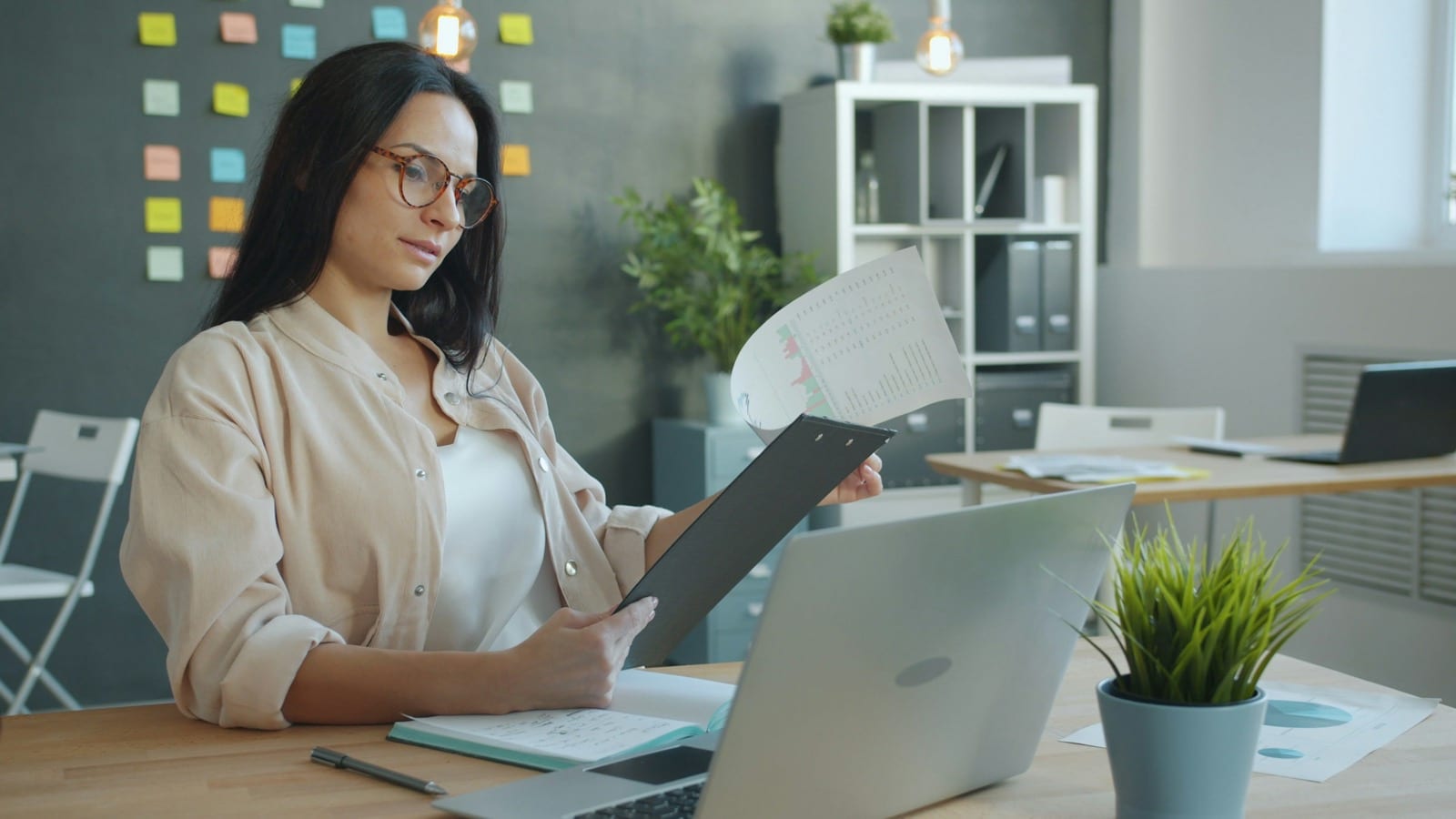 A person in glasses reads papers on a clipboard, seated at a laptop.