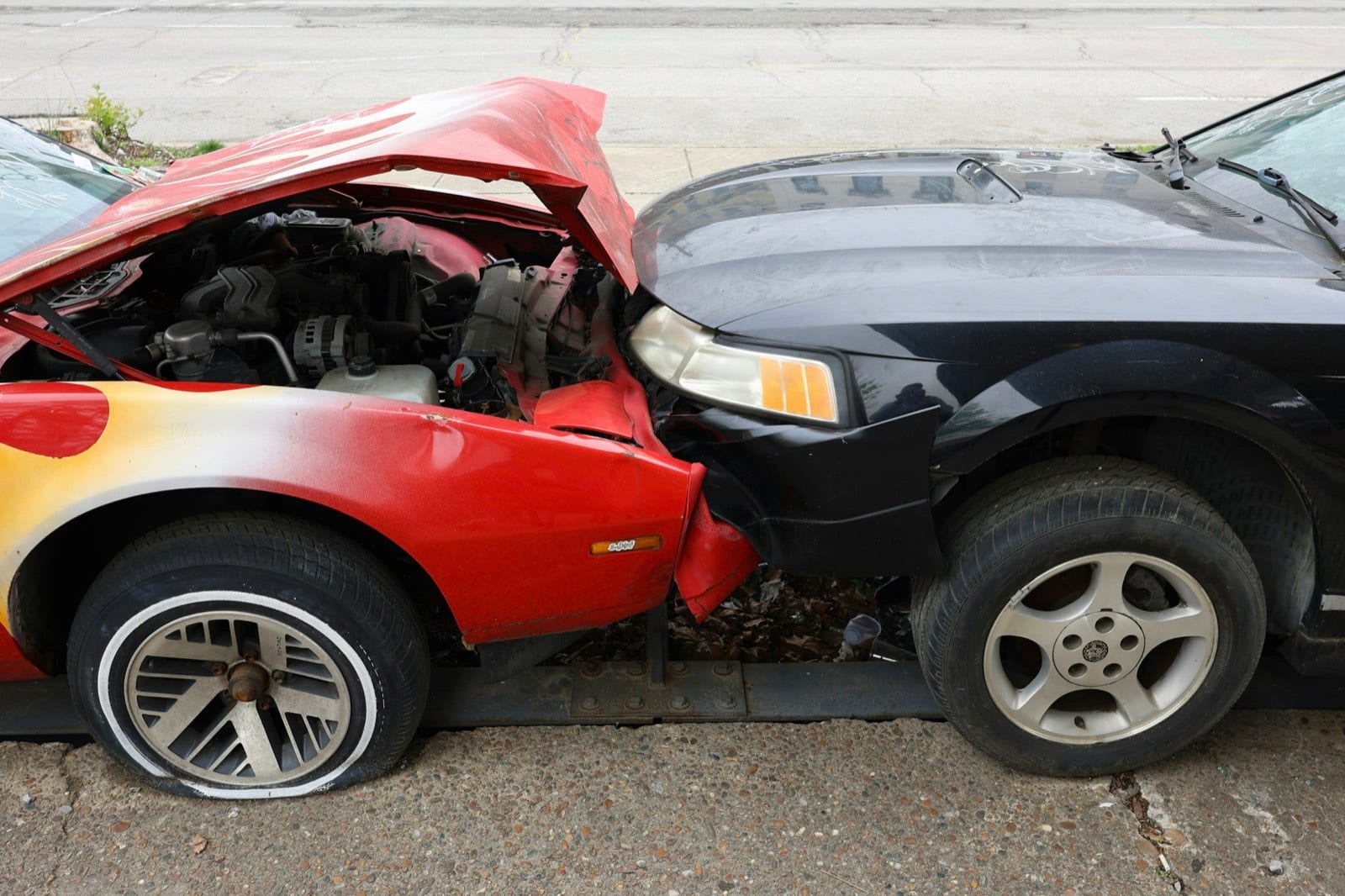 Two cars, one red and one black, with their fronts crashed together; the red car’s hood is crumpled and raised.